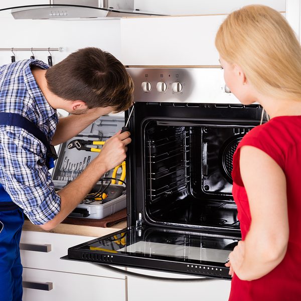Male Worker Repairing Oven Appliance In Presence Of Young Woman In Kitchen Room